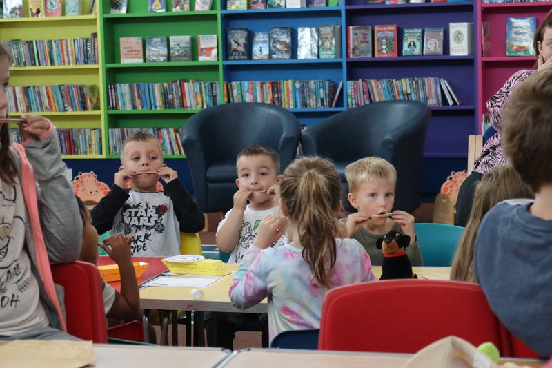 Children sit around a table in a library taking part in a music-themed activity, with colourful bookshelves in the background.