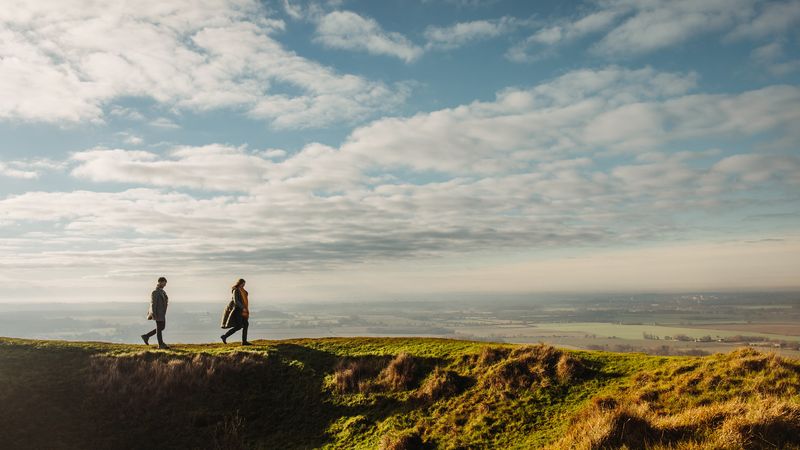 Two people walking along a grassy ridge with panoramic views of the Kent countryside beneath a wide sky filled with clouds.