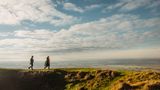 Two people walking along a grassy ridge with panoramic views of the Kent countryside beneath a wide sky filled with clouds.