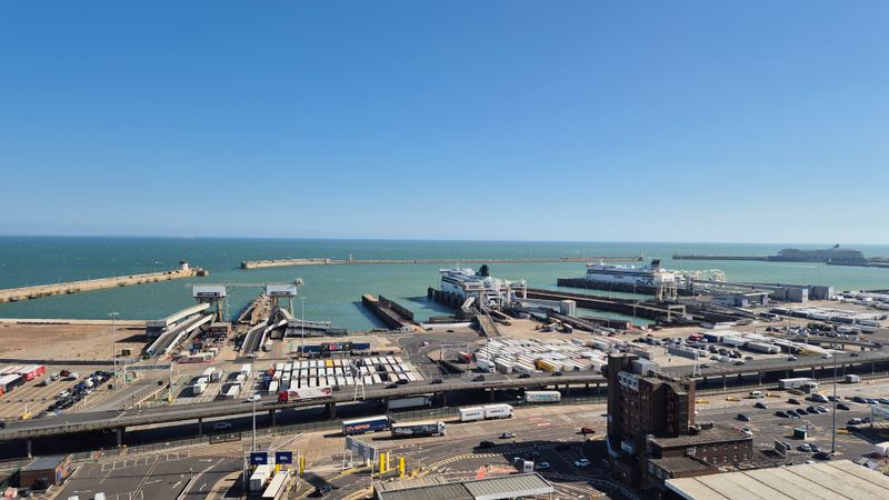 A panoramic view of the Port of Dover showing ferry berths, terminal facilities, roads, freight traffic and ferries in the harbour, with the English Channel extending to the horizon under clear skies.