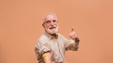 Older man against orange background with shirt sleeves rolled up and animal-themed plaster on his arm gives thumbs up