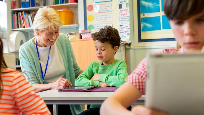 An older woman sits down next to a child sitting at a desk to help them with their work. They are both smiling and wearing green.