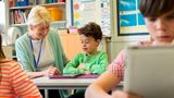 An older woman sits down next to a child sitting at a desk to help them with their work. They are both smiling and wearing green.