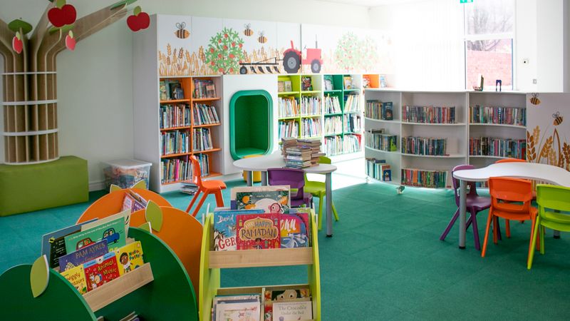 &ldquo;A colourful children&rsquo;s library area with bright green carpet, low bookshelves filled with books, and playful tree and farm-themed wall decorations. Small tables and chairs in various colours are arranged in the centre.&rdquo;
