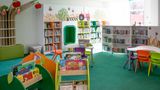 &ldquo;A colourful children&rsquo;s library area with bright green carpet, low bookshelves filled with books, and playful tree and farm-themed wall decorations. Small tables and chairs in various colours are arranged in the centre.&rdquo;