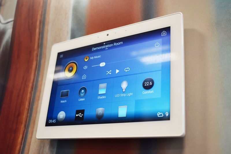 Wall-mounted touchscreen smart home control panel showing music playback, lighting, shades, and temperature settings on display