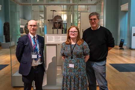 Three people standing in a museum gallery beside a glass display case containing an ornate Anglo-Saxon helmet, with exhibit panels and artefacts visible in the background.