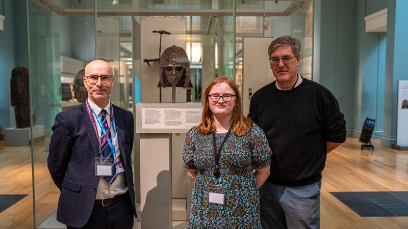 Three people standing in a museum gallery beside a glass display case containing an ornate Anglo-Saxon helmet, with exhibit panels and artefacts visible in the background.