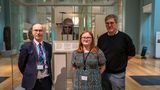 Three people standing in a museum gallery beside a glass display case containing an ornate Anglo-Saxon helmet, with exhibit panels and artefacts visible in the background.