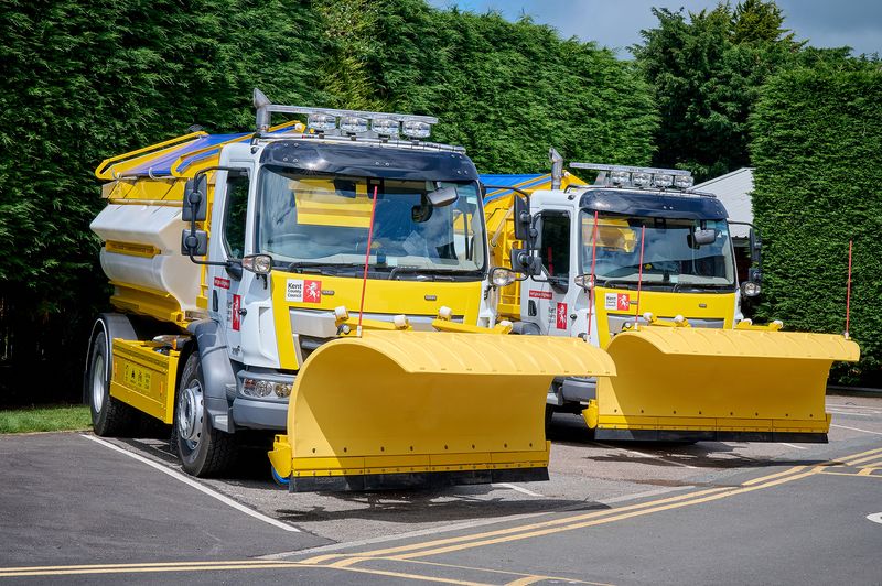 Two yellow and white Kent County Council gritter vehicles fitted with large snowplough blades are parked side by side in a depot car park, with tall green hedges and trees in the background. The vehicles display KCC branding and are equipped for winter road maintenance.