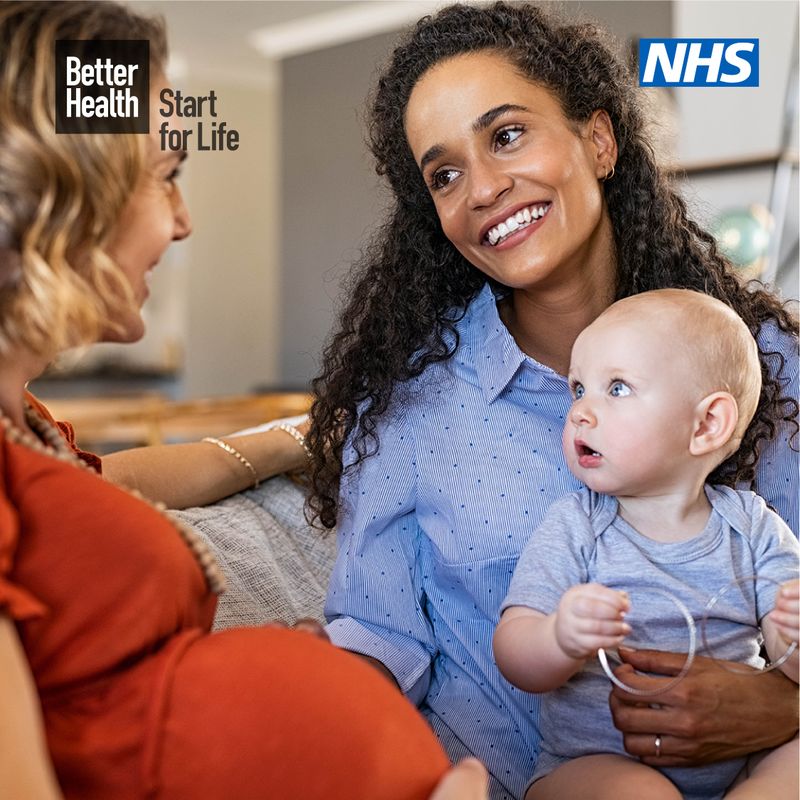 Two women - one pregnant and one nursing a young baby - sat on a grey sofa 