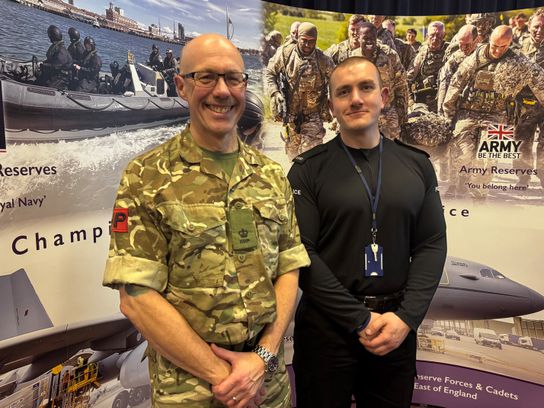 Two Kent reservists standing in front of a reservists recruitment banner. One is in camouflage gear and the other is in a black police uniform