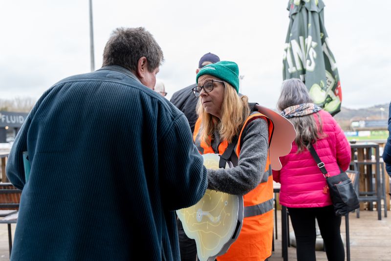 A stop smoking adviser wearing a hi-vis jacket and green woolly hat speaking to a football fan - tables and chairs in the foreground and the pitch is in the distance