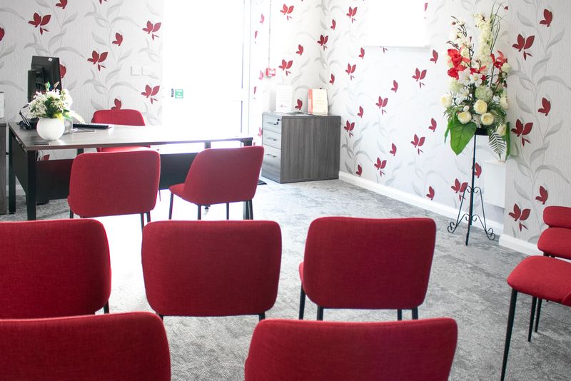 A small ceremony-style room with rows of red chairs facing a desk at the front. The walls have a white and red floral pattern, and a tall arrangement of flowers stands on a metal stand to the right