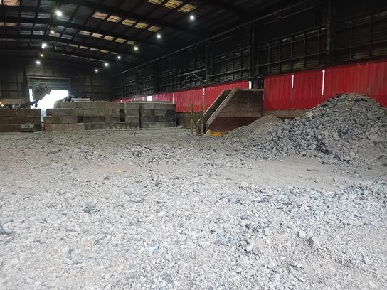 Interior of a waste transfer station during refurbishment, showing a rough, excavated floor with piles of broken concrete and rubble, partially dismantled bay walls, and red safety sheeting along the side.