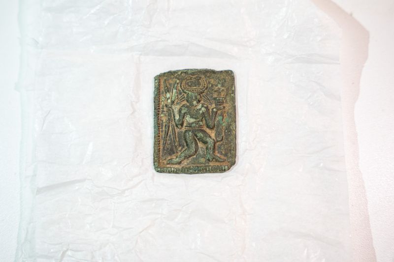 Small rectangular metal artefact with a corroded green surface, depicting a seated figure in relief, placed on white conservation material.