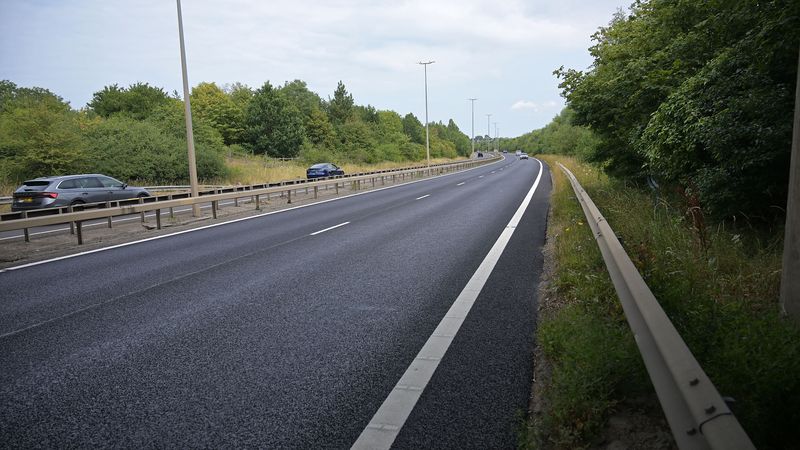 Freshly resurfaced carriageway on the A299 Thanet Way, showing a smooth black road surface, central barrier and street lighting, with trees and vegetation lining both sides of the dual carriageway.