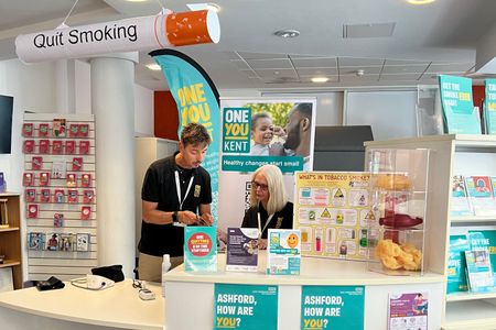 Two staff at the information stand at the Ashford One You Kent shop that provides quit smoking advice. The display includes stop smoking leaflets, a &ldquo;Quit Smoking&rdquo; ceiling decoration, a poster showing harmful chemicals in tobacco smoke, and a model display illustrating the effects of smoking.