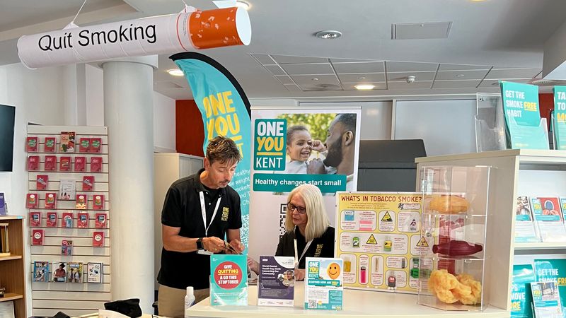 Two staff at the information stand at the Ashford One You Kent shop that provides quit smoking advice. The display includes stop smoking leaflets, a &ldquo;Quit Smoking&rdquo; ceiling decoration, a poster showing harmful chemicals in tobacco smoke, and a model display illustrating the effects of smoking.