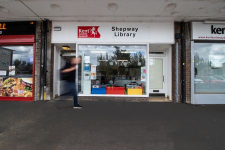 The outside of a Shepway Library - it is a building with a big wide window and the words 'Shepway library' at the top. There is a blurry figure walking past.