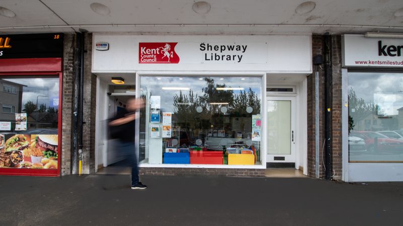 The outside of a Shepway Library - it is a building with a big wide window and the words 'Shepway library' at the top. There is a blurry figure walking past.
