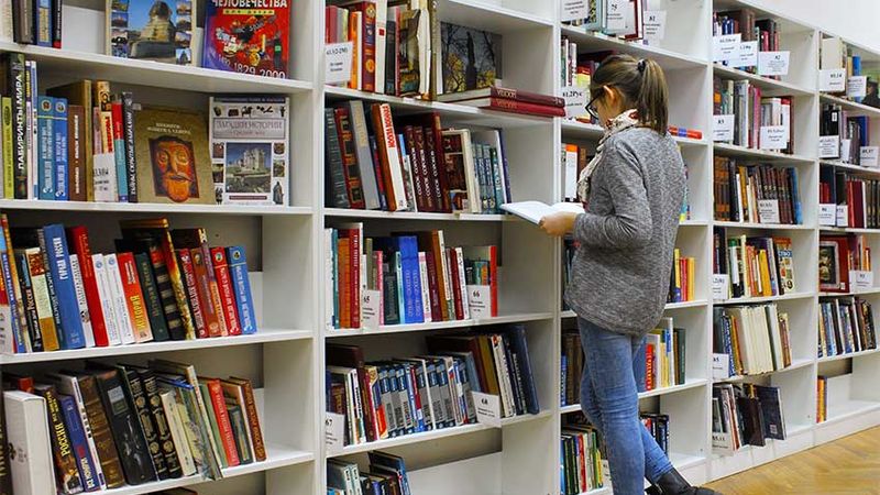 Image of a woman standing infront of shelves of colourful library books, reading a book
