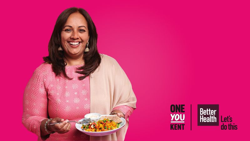 An overweight woman in a pink saree holding a plate of food consisting of lentils, yoghurt, and a pitta bread. 