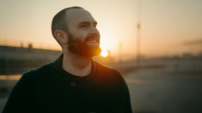 Liam Cumber stands holding a takeaway coffee cup, backlit by the sun, looking to the left. He has a beard and is wearing a dark woollen jumping. 