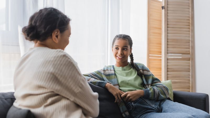 Smiling teenage girl talking with her mother while sat at on the sofa at home 