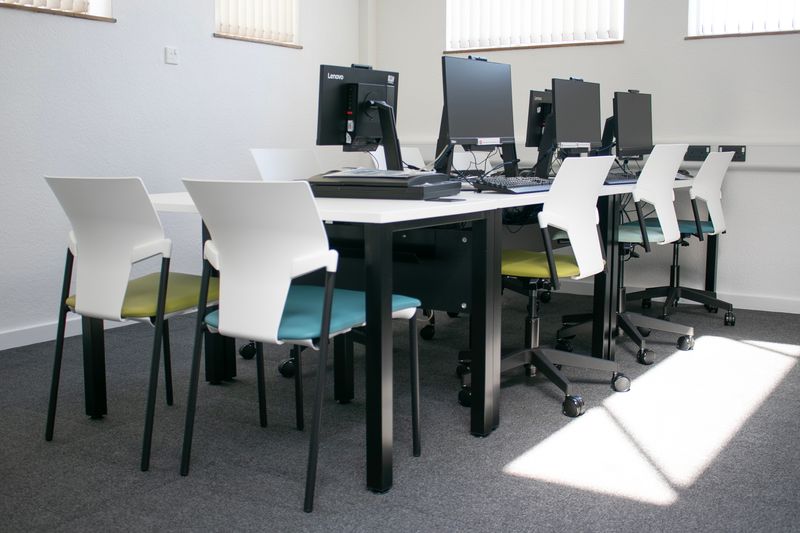 A bright computer room with two rows of desks holding desktop computers. White chairs with coloured seats are arranged around the tables, and sunlight falls across the grey‑carpeted floor.