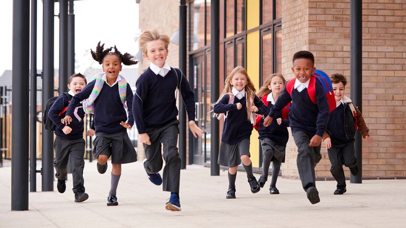 A group of primary school&ndash;aged children wearing school uniforms and backpacks run toward the camera along a covered walkway outside a brick school building.