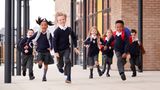 A group of primary school&ndash;aged children wearing school uniforms and backpacks run toward the camera along a covered walkway outside a brick school building.
