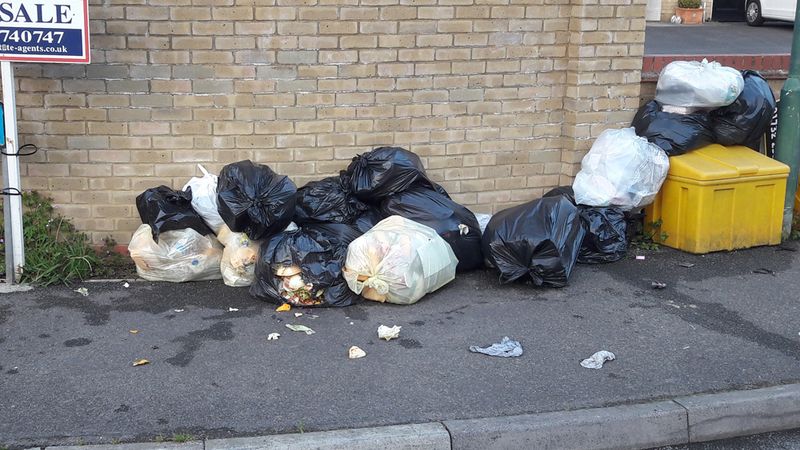 Black and white rubbish bags piled on a pavement against a brick wall, with loose food waste spilling from some bags, next to a yellow bin and a &ldquo;For Sale&rdquo; sign; litter is scattered around the kerb.