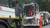 Road resurfacing work on a two‑lane rural road: a paving machine spreads fresh asphalt while a large truck supplies material, and a road worker in high-visibility orange clothing and a hard hat stands beside the machine guiding the operation, with trees lining the roadside.