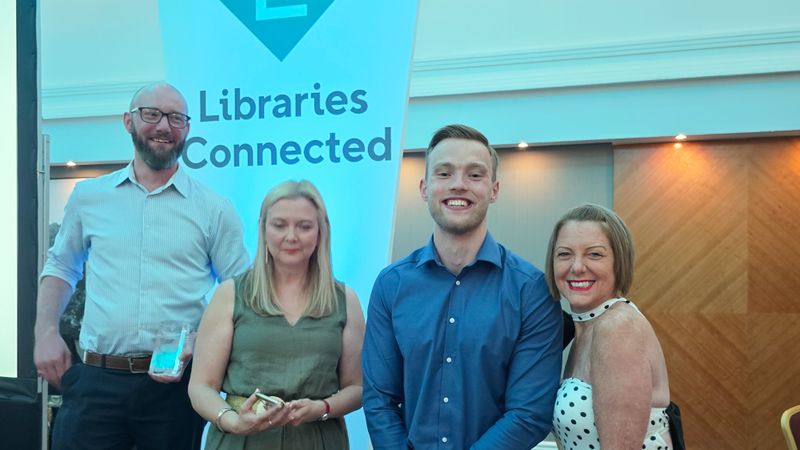 Four people posing beside a &ldquo;Libraries Connected&rdquo; banner at an indoor awards event. One person holds a glass award while the group stands in front of wood-panelled walls and conference-style lighting.