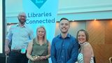 Four people posing beside a &ldquo;Libraries Connected&rdquo; banner at an indoor awards event. One person holds a glass award while the group stands in front of wood-panelled walls and conference-style lighting.