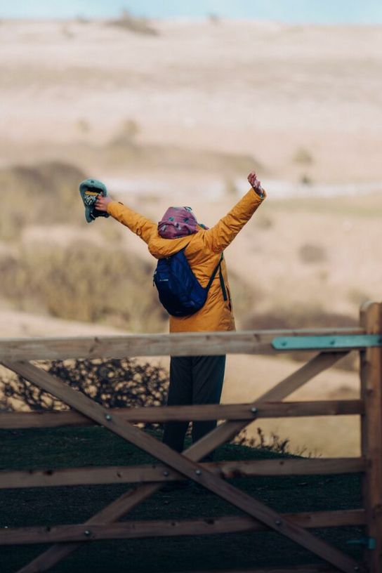 A walker wearing a backpack stands beside a wooden gate in the countryside with open downland stretching into the distance.
