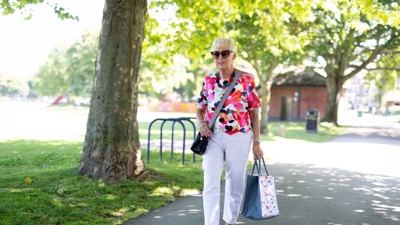 Person walking along a tree-lined park path carrying shopping bags, wearing a colourful floral top and sunglasses on a sunny day.