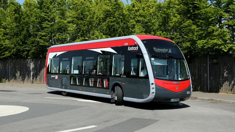 A red and grey Fastrack electric bus travels along a road on a bright sunny day. The modern single-deck bus displays "Bluewater A" on the destination screen and is surrounded by tall green trees and a wooden fence in the background. Visible branding on the vehicle includes "fastrack electric."