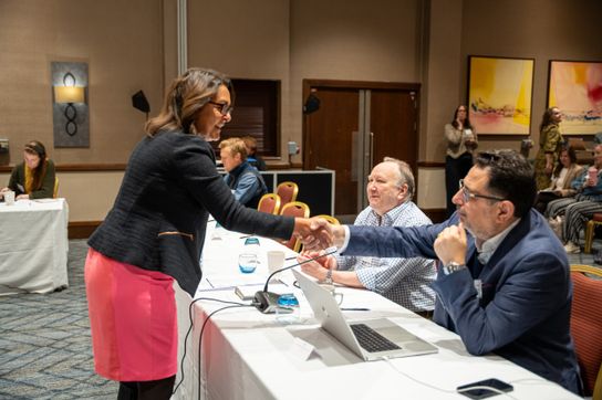 A participant greets a member of a panel with a handshake across a conference table during a Kent water sector meeting. Other attendees sit nearby with laptops, microphones and papers in a hotel conference room.