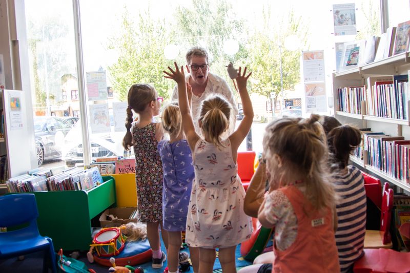 A group of children in a library with their arms raised. There is a woman facing them with a big smile on her face.