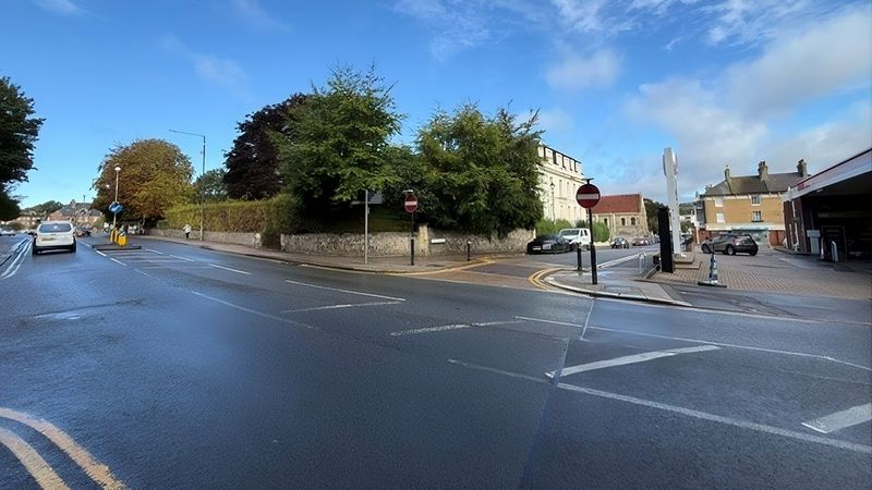 Wide view of a road junction near Dover Priory, showing a wet roadway, traffic islands and road signs. A stone wall and mature trees surround a large light-coloured building in the centre background, with a church-like building visible behind it. Several cars are parked or travelling nearby, and a bright blue sky with scattered clouds stretches overhead.