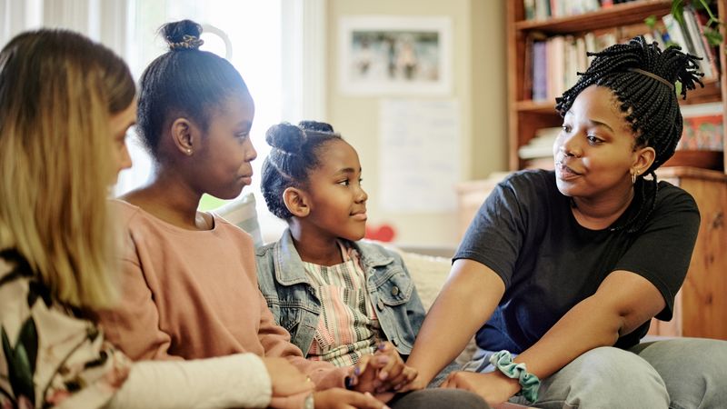 A mum and two daughters sit on a sofa and get support from another adult female