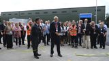 The image shows a group of people clapping as KCC's Chairman, Bryan Sweetland, cuts a green ribbon to showcase the opening of the new Waste Transfer Station. The group of people stand in front of a big dark green warehouse which is the new facility. 