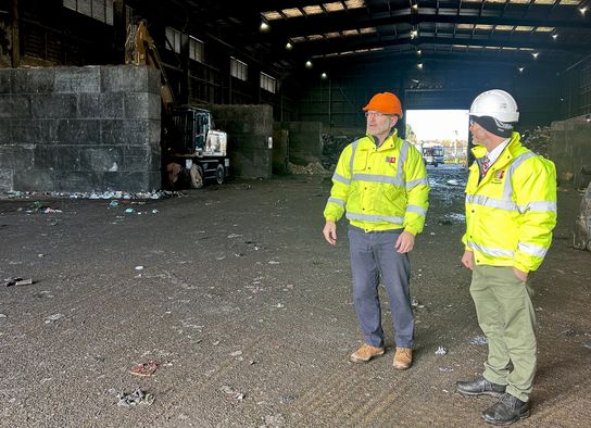 Two men in hi viz stand in a big waste transfer station.