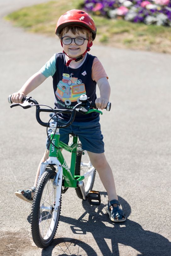 Young child wearing a red cycle helmet and glasses riding a small green bicycle on a smooth park path, with flowers visible in the background.