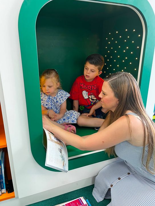A mum reads a book with her two young children seated inside a green reading pod. The children are looking at the open book while sitting together in the enclosed space, which has small lights on the back wall. Bookshelves and library materials are visible nearby.