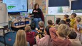 Children sitting on the carpet in their school classroom. They are joining in with a teacher who is reading a story in Makaton at the front of class