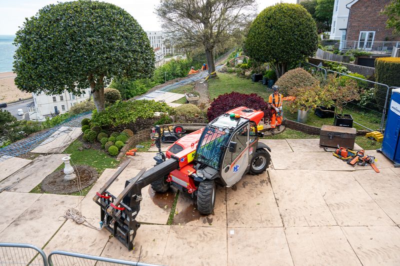 Construction site on a terraced garden overlooking the coast, with a construction equipment in the foreground and workers setting up slope stabilisation netting down the hillside.