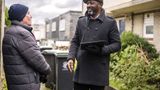 Two men talk on a doorstep. One is older than the other. One holds a clipboard